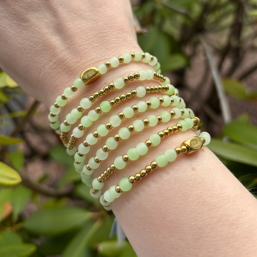 Hand wearing multiple green and gold beaded bracelets with a natural background