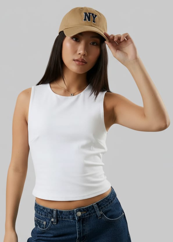 Woman wearing a white tank top, blue jeans, and a blue cap against a beige background
