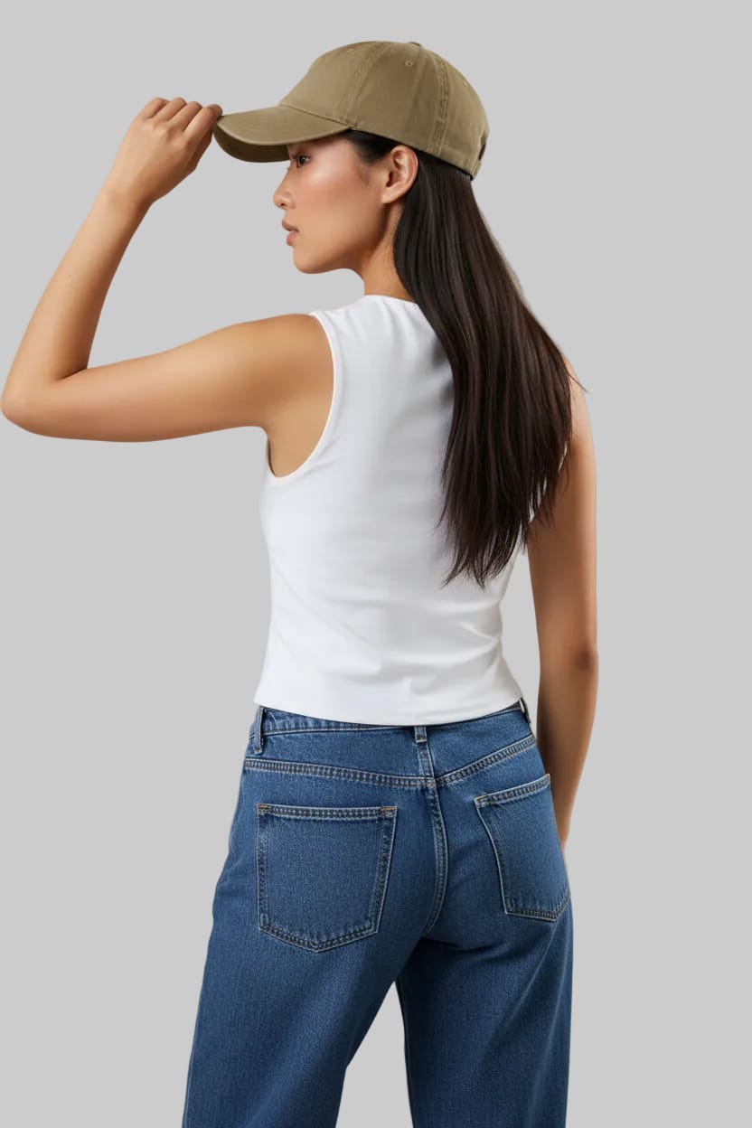 Woman wearing a white sleeveless top and blue jeans, adjusting a blue cap on a beige background