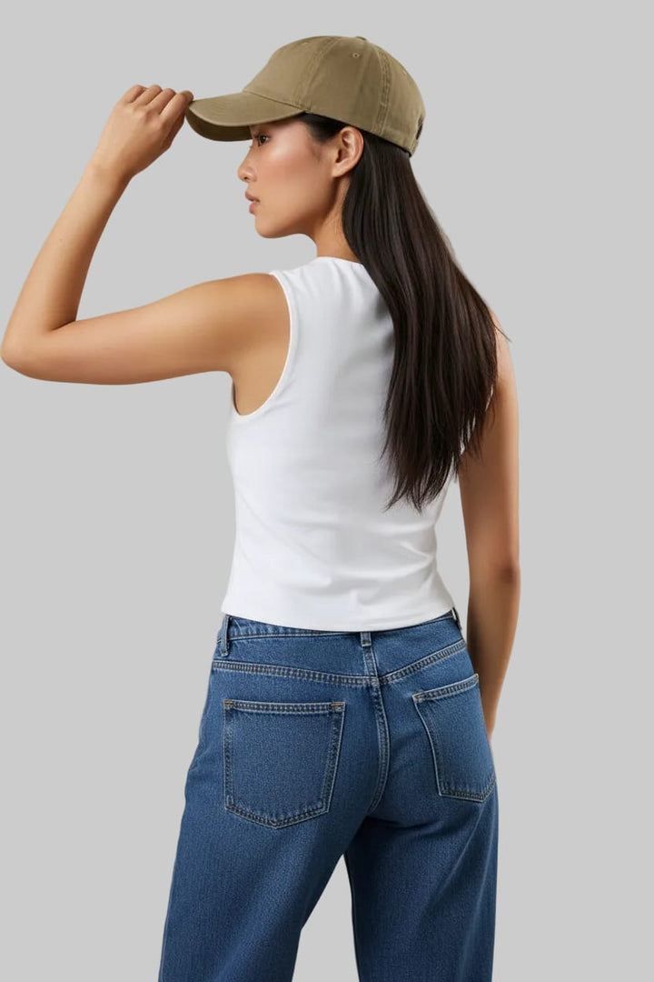 Woman wearing a white sleeveless top and blue jeans, adjusting a blue cap on a beige background